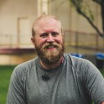 A cheerful bearded man smiling outdoors in a casual setting with greenery.